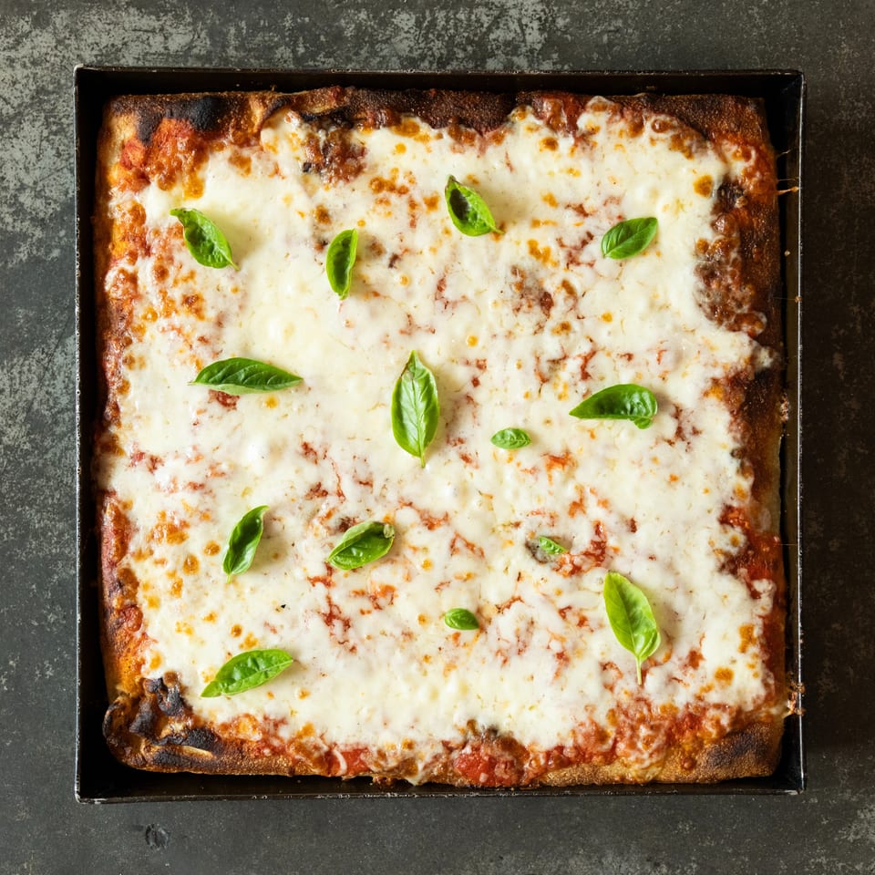 Overhead view of a rectangular, cheese-covered pizza topped with tomato sauce and fresh basil in a black baking tray.