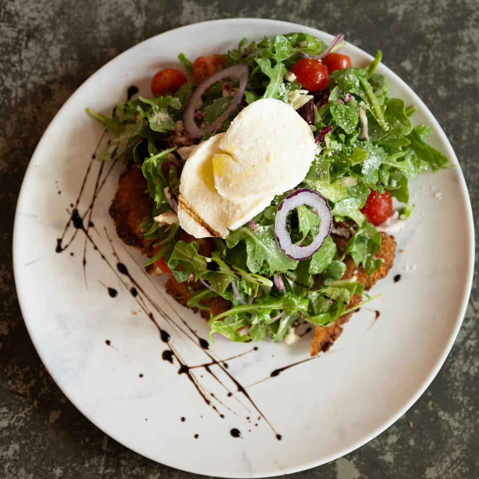 Breaded entrée topped with arugula salad, cherry tomatoes, red onion, soft cheese, and balsamic drizzle on a white plate.