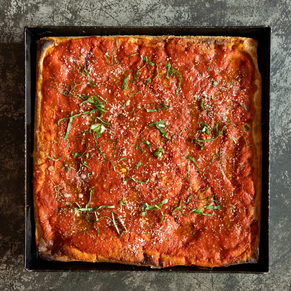 Rectangular pizza topped with tomato sauce, herbs, grated cheese, and basil in a black baking tray.