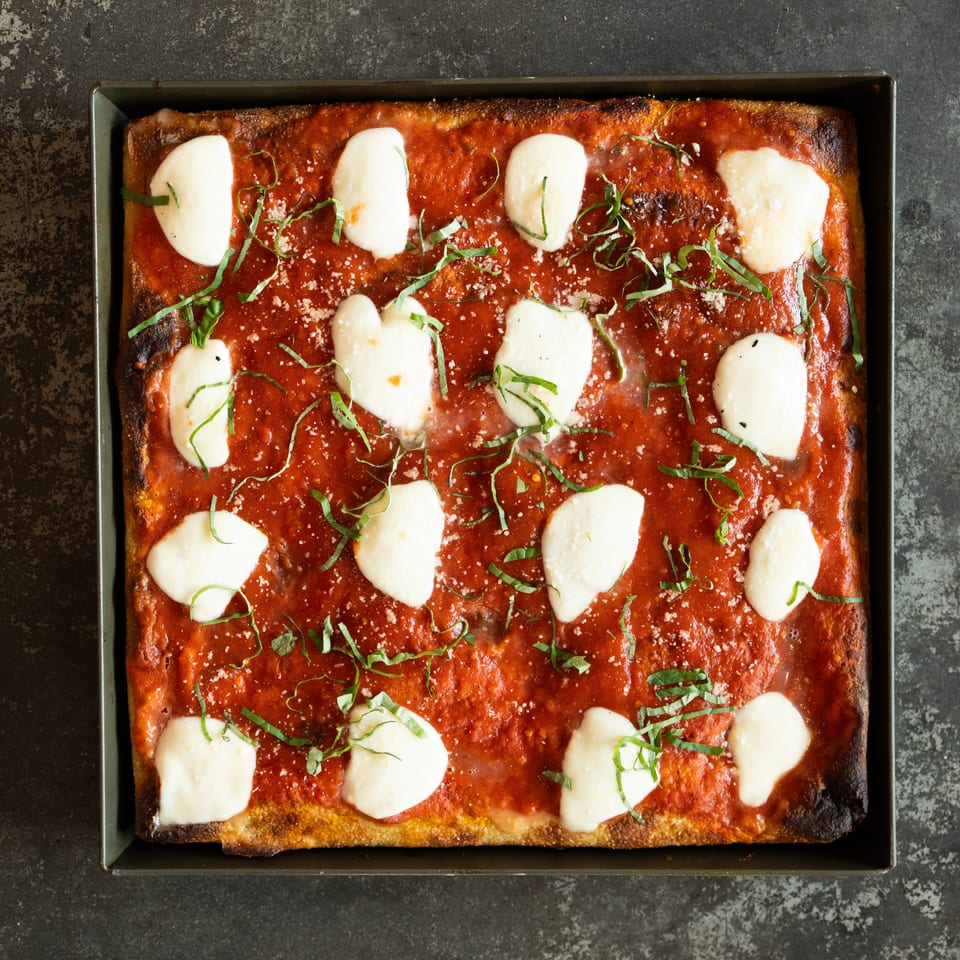 Rectangular tomato pizza topped with fresh mozzarella, basil, grated cheese, and charred crust in a baking tray.