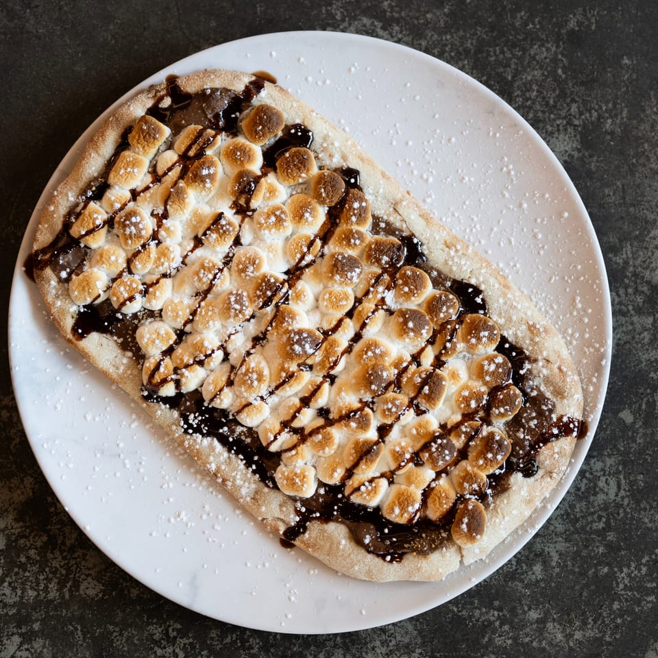 Dessert pizza topped with toasted marshmallows, chocolate drizzle, and powdered sugar on a white plate.