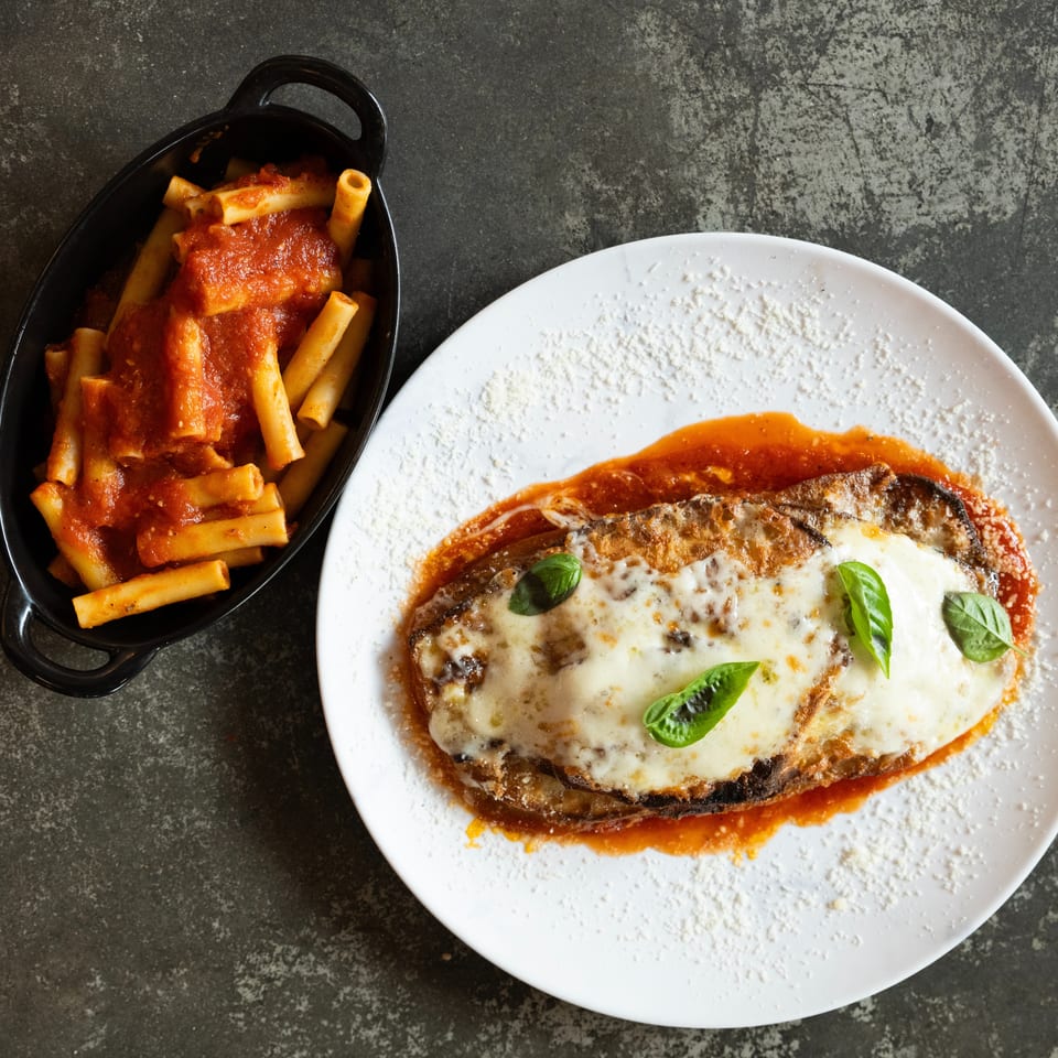 Eggplant Parmesan topped with melted cheese and basil, served with tomato-sauced ziti in a black baking dish.