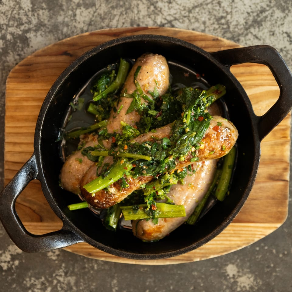 Roasted sausages topped with sautéed leafy greens and chili flakes in a cast-iron skillet on a wooden board.