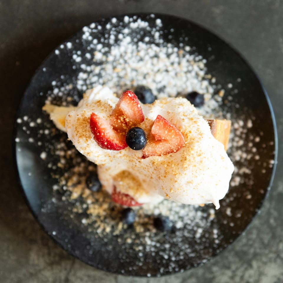 Whipped cream dessert topped with sliced strawberries and blueberries on a black plate dusted with powdered sugar.