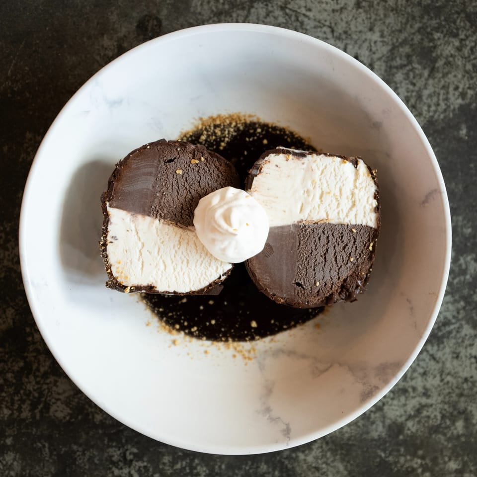 Chocolate and vanilla ice cream cake slices topped with whipped cream on a white plate with chocolate sauce.