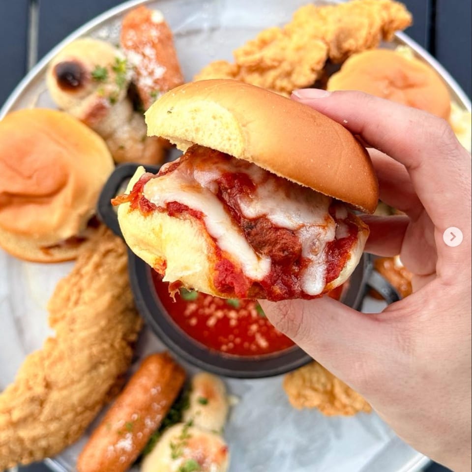 Hand holding a cheesy meatball slider over a platter of appetizers and marinara sauce.