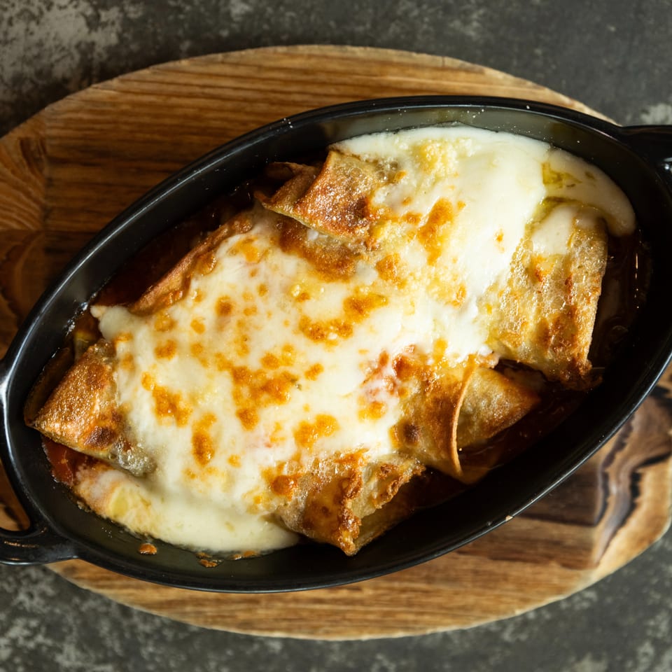 Baked pasta squares topped with melted, browned cheese in a black oval casserole dish on a wooden board.