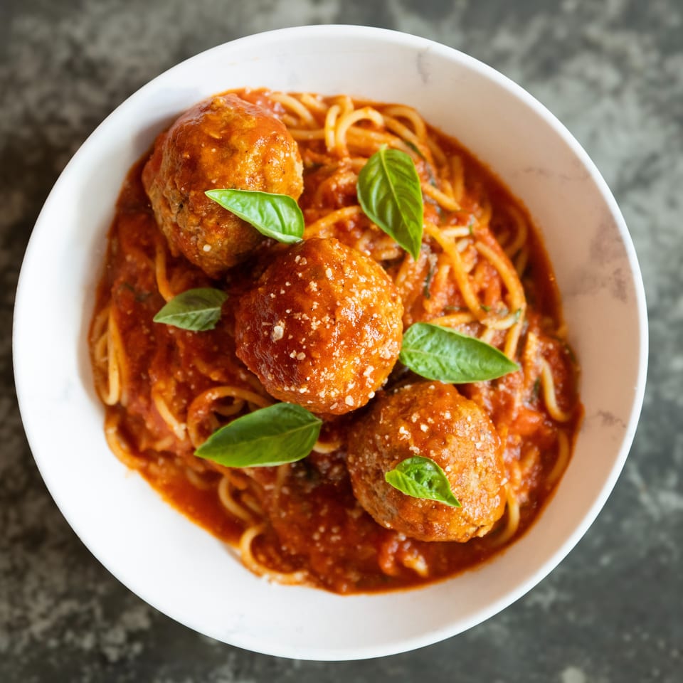 Bowl of spaghetti and meatballs in tomato sauce, garnished with fresh basil and grated cheese.